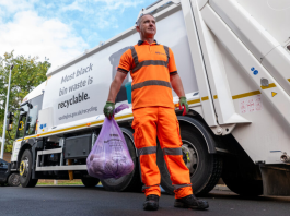 Photo of man standing on a road in orange high vs overalls holding a full waste sack with a waste lorry in the background.