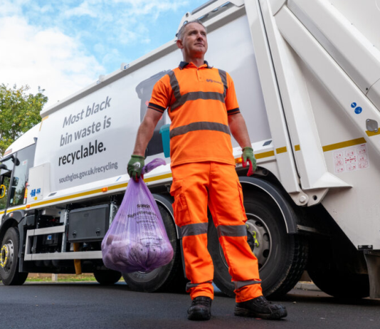 Photo of man standing on a road in orange high vs overalls holding a full waste sack with a waste lorry in the background.
