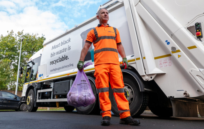 Photo of man standing on a road in orange high vs overalls holding a full waste sack with a waste lorry in the background.